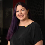 Image is a headshot of a femme-presenting person with long black-purple hair and wearing a black scoop neck tshirt. She is looking at the camera and has a soft smile.
