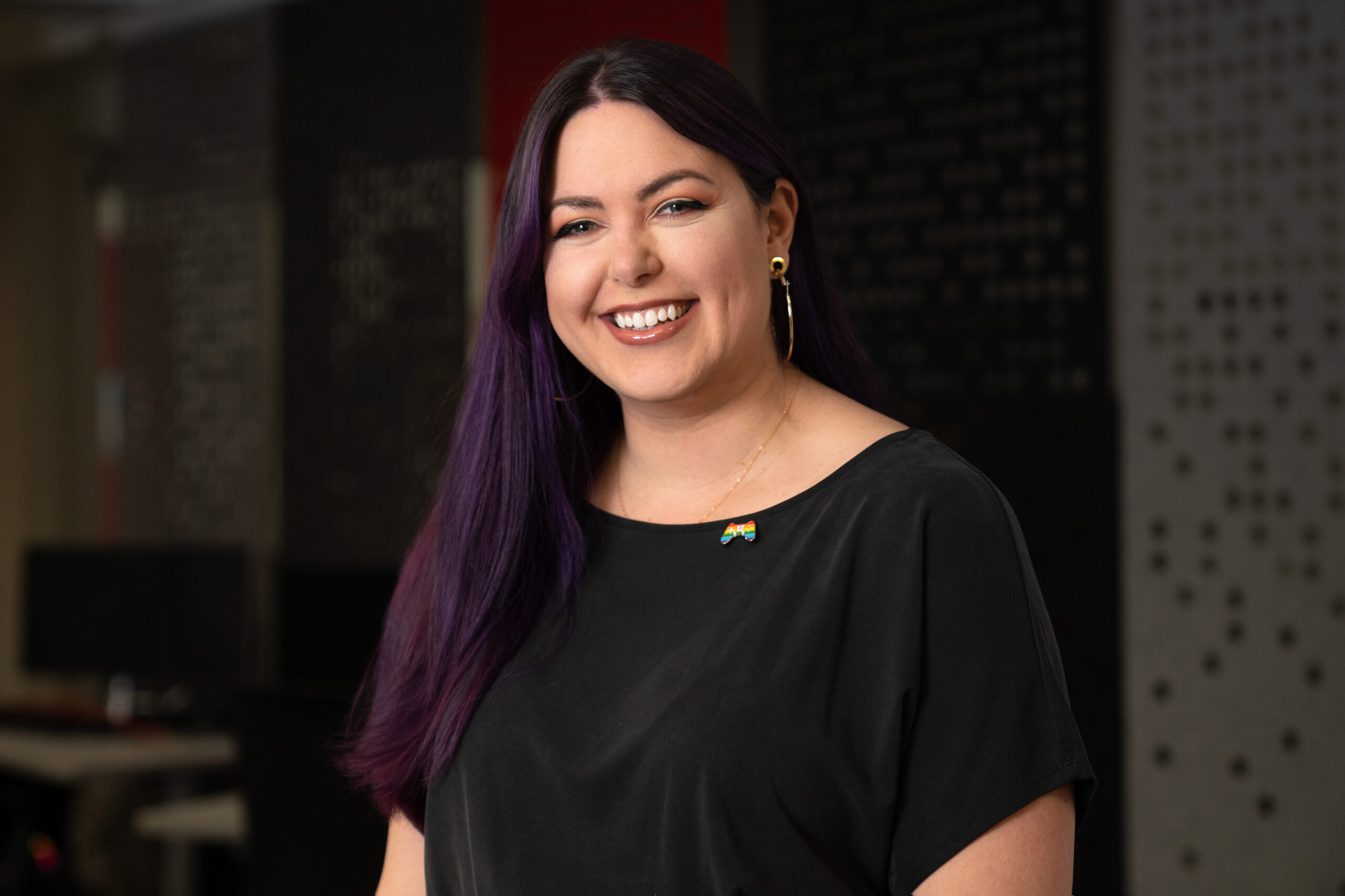 Image is a headshot of a femme-presenting person with long black-purple hair and wearing a black scoop neck tshirt. She is looking at the camera and has a soft smile.