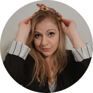 Circular image with a white woman in the center. She has blonde hair topped with a colorful headband, a quirky smile, and wearing a black blazer with white cuffs.