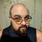 Headshot of a man with a shaved head and goatee and who is wearing orange-tinted glasses