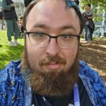 Headshot of a white masc-presenting person with brown hair, beard, and mustache. They are wearing glasses anda blue print button up over a graphic tee shirt