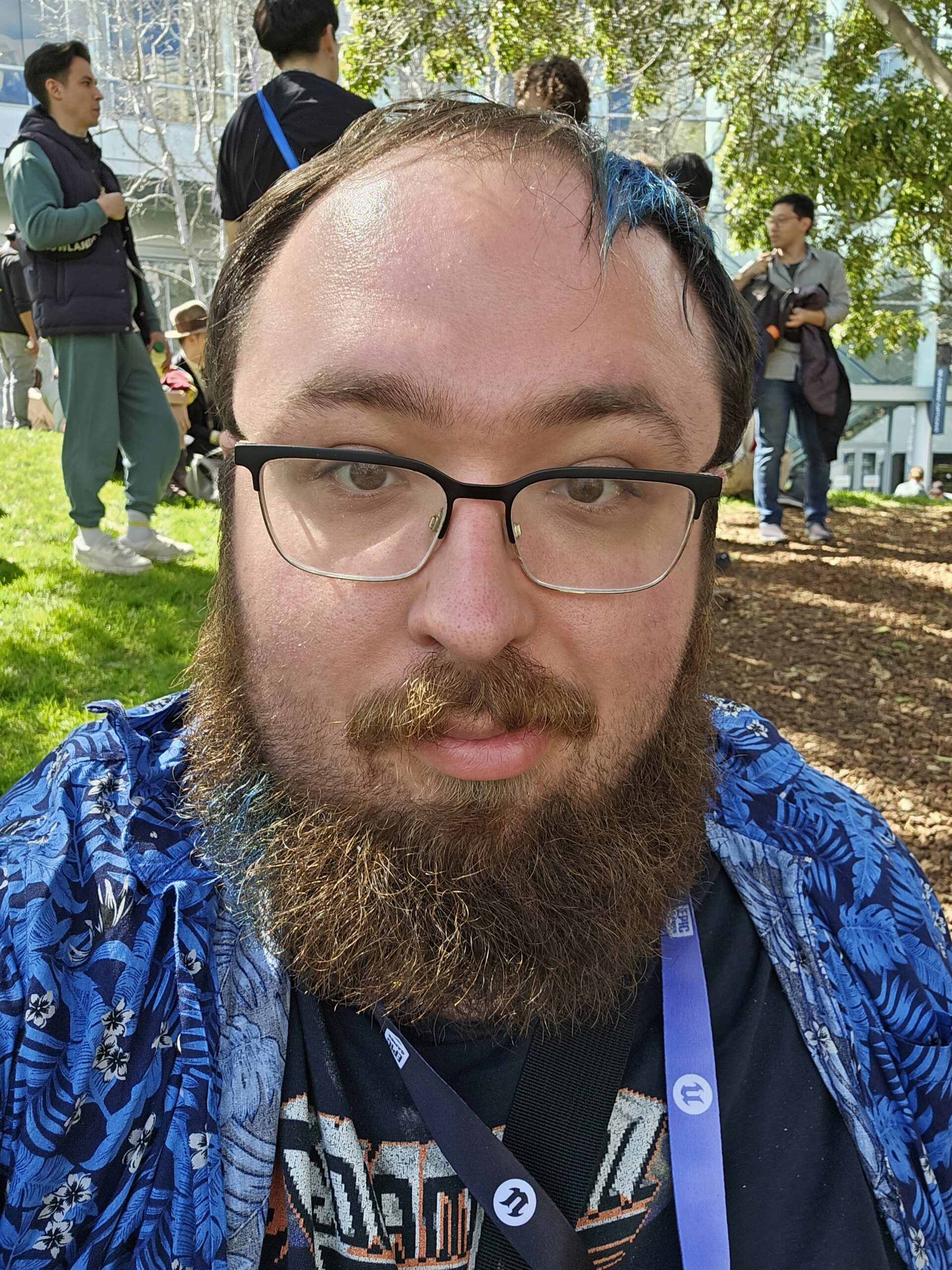 Headshot of a white masc-presenting person with brown hair, beard, and mustache. They are wearing glasses anda blue print button up over a graphic tee shirt