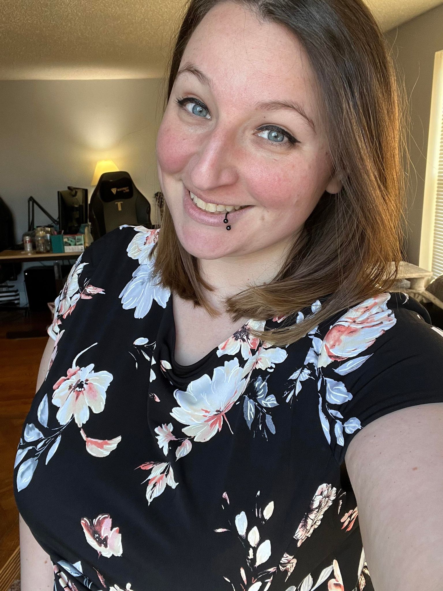 Image of a femme-presenting person from the waist up. They have blonde hair, soft eyes, and a quirky smile. They are wearing a black top with a floral print