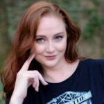 Headshot of a woman with red hair and fair skin. She has a gentle face and is smiling slightly with one hand up by her face.