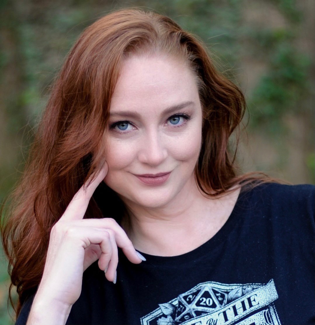 Headshot of a woman with red hair and fair skin. She has a gentle face and is smiling slightly with one hand up by her face.
