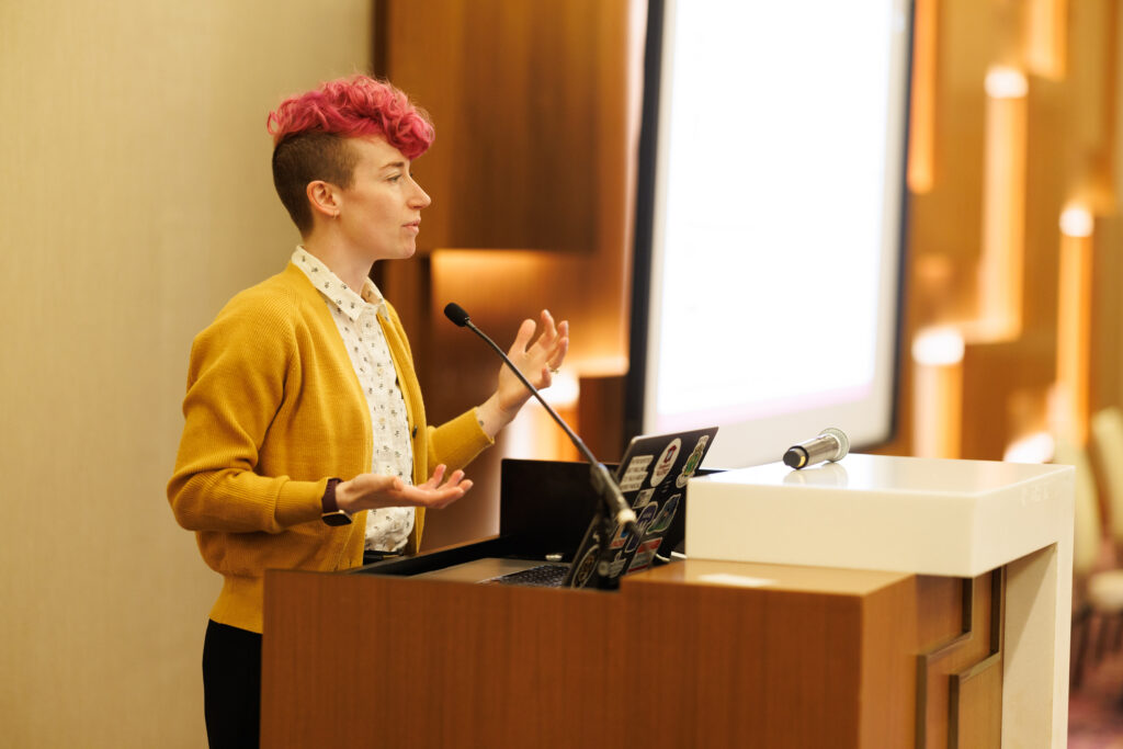 Dr. Elizabeth Kilmer stands behind a podium and microphone in a conference room giving a workshop at the DICE 2025 event.