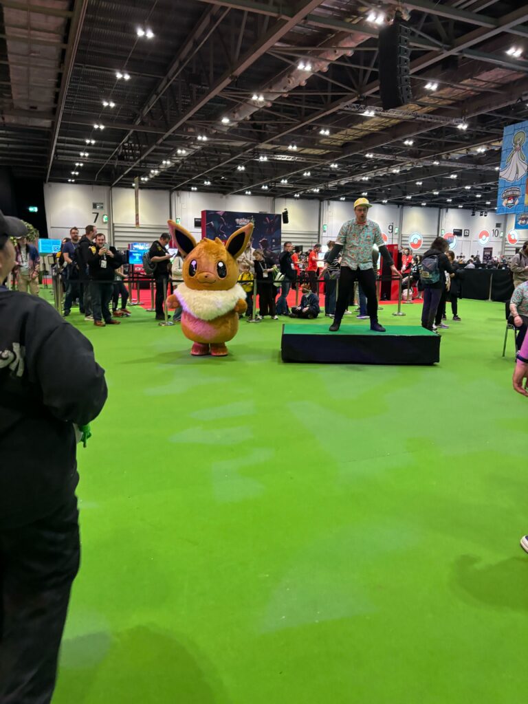 green turf flooring with a large evee mascot and human pokemon trainer on a pedestal leading yoga on the convention floor.