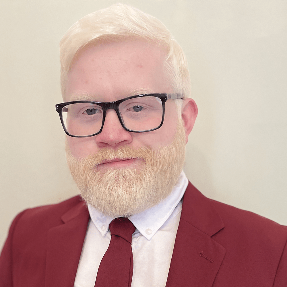 Headshot of a bearded albino male-presenting person with black glasses, red suit jacket and tie, and a white shirt. 