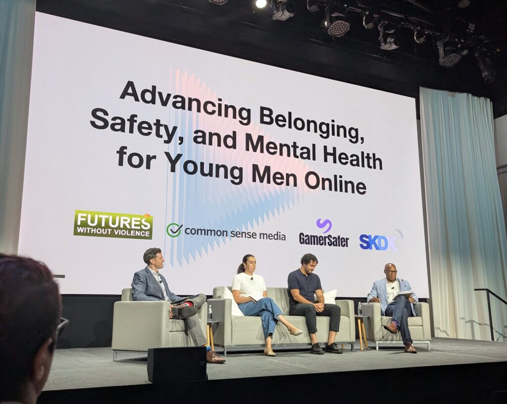 Four individuals (Michael Frazier, Maria Tamellini, Michael Robb, and Jesse Mahler) sit in chairs on a stage during a panel in front of a screen. On the screen is text that reads "Advancing Belonging, Safety, and Mental Health for Young Men Online."