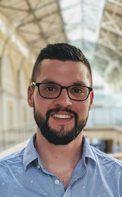 A headshot of Brian Kunde, a man wearing a blue button up shirt with glasses, short dark hair, and a beard.