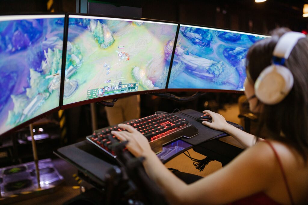 A photo of a person playing video games at a pc setup. They're wearing headphones, and using a keyboard and mouse to play a game across three monitors